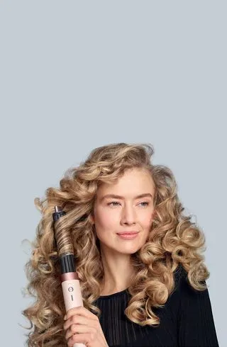 Жінка тримає в руці рожево-золотистий мультистайлер Dyson Airwrap Ceramic Pink. Довге пасмо волосся автоматично накручується навколо довгої циліндричної насадки завдяки сильному потоку повітря (ефекту Коанда). Видно сформований пружний локон